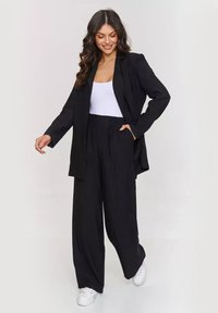 Czarna marynarka z długimi rękawami i luźnym krojem, zestawiona z szerokimi czarnymi spodniami i białym topem na ramiączkach; modelka nosi białe sneakersy.