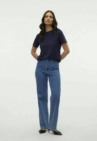 Tume sinise kaelusega T-särk, mis on kombineeritud kõrge vöökohtaga helesiniste teksadega. Mudel seisab enesekindlalt, käed puusadel, jalas mustad terava ninaga kontsad.