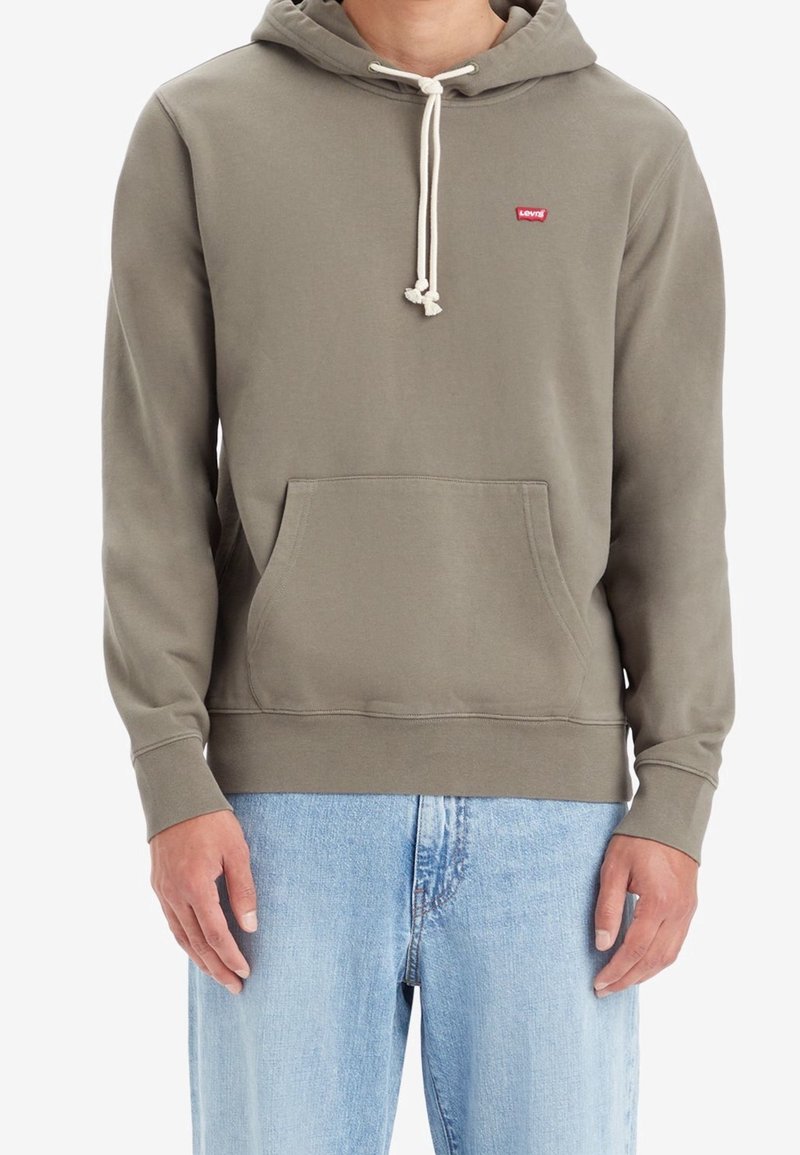 Homme portant un sweat à capuche Levi's vert olive avec poche avant et cordons blancs, associé à un jean bleu clair, debout devant un fond blanc.