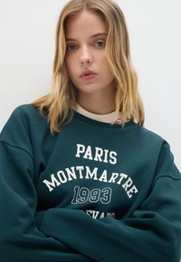 Sudadera verde oscuro con texto gráfico blanco "PARIS MONTMARTRE 1993" en el pecho. Cuello acanalado y textura suave.