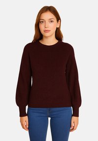 Kvinna med rakt brunt hår klädd i en mörk vinröd långärmad tröja och blå jeans, stående mot en ljus, enfärgad bakgrund.