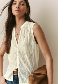 Blouse sans manches crème avec un motif en dentelle texturée, devant à boutons, col mandarin, associée à une ceinture tissée beige et un sac à main en suède marron.