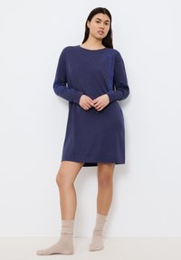 Nightdress lungo con maniche, che arriva al ginocchio, in tessuto blu scuro con ricami floreali azzurri lungo il lato, indossato con calzini leggeri a coste.