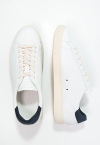 Baskets en cuir blanc avec une semelle en caoutchouc crème, avec un accent talon bleu marine, des lacets beige, et un design minimaliste.