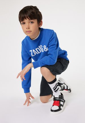 Sweatshirt bleu avec texte blanc, shorts noirs, baskets noir et blanc avec accents rouges, et chaussettes rayées ; pose à genoux.