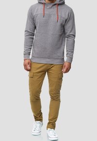 Grijze hoodie met oranje trekkoorden, voorzien van een structuurstof en diagonale naaddetails. Gecombineerd met beige cargobroek en witte sneakers.