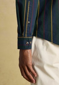 Detailný záber na tmavú ruku vedľa manžety dlhého rukáva trička farieb námorníckej modrej, žltej a červenej, položeného na bielych nohaviciach.