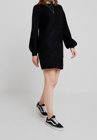 Robe en maille noire avec de longues manches bouffantes ; poignets et ourlet côtelés. Associée à des baskets noir et blanc avec des lacets classiques et des rayures sur les côtés.