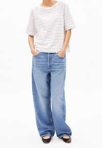 Persoon in een wit shirt met korte mouwen met strepen, losse lichtblauwe jeans met handen in de zakken, en zwarte platte schoenen tegen een witte achtergrond.