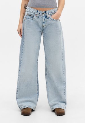 Personne portant un jean large bleu clair, un débardeur gris court, des chaussures marron, avec une main dans la poche et un tatouage visible sur le bras.