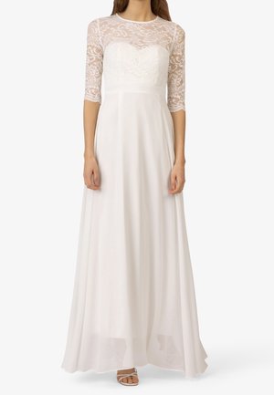 Femme portant une longue robe blanche avec un corsage en dentelle et des manches trois-quarts en dentelle, associée à des sandales blanches à bout ouvert.