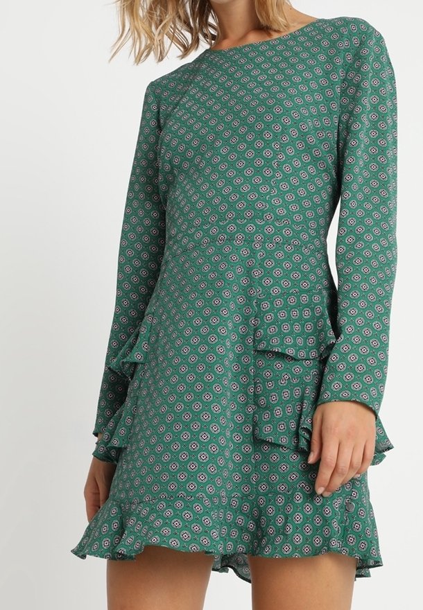 Vihreä mekko, jossa on pitkät hihat, röyhelöinen helma ja kerroksellinen muotoilu. Sisältää geometrisen kuvion vaaleanpunaisena ja valkoisena teksturoidulla kankaalla.