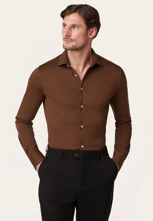 Man in een bruin overhemd met lange mouwen en zwarte broek, handen in de zakken, kijkt opzij tegen een effen lichte achtergrond.
