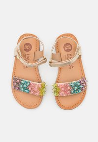 Sandalias de cuentas con una tira frontal multicolor que presenta cuentas rosas, azules, verdes y moradas. Planta de cuero marrón y tira metálica.