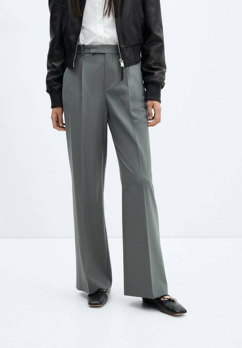 Personne portant un pantalon gris taille haute à jambes larges avec des plis sur le devant, une veste en cuir noire, une chemise blanche et des ballerines noires à bout pointu.