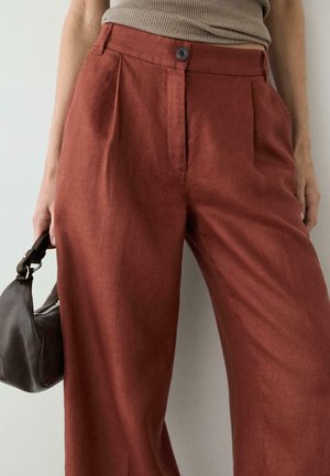 Femme portant un pantalon large taille haute couleur rouille, top côtelé beige, tenant un petit sac à main en cuir marron foncé à côté.