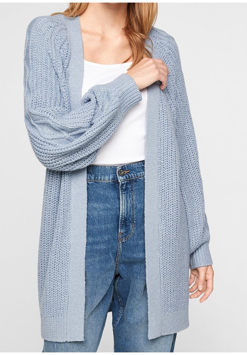Cardigan bleu clair en maille avec ouverture sur le devant, poignets côtelés et motifs texturés. Porté sur un haut blanc et associé à un jean en denim.