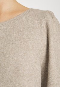 Beige stickad tröja med vid halsringning och subtil textur, med något puffärmar och ribbat detaljer vid kragen.