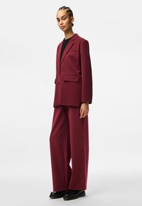 Burgundi kohandatud ülikond kahekordse pealsega ja laia säärega pükstega. Sile kangas, lõikeklappid ja ees olevad taskud. Musta värvi jalanõud.