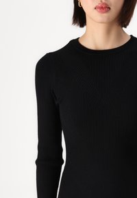 Pull noir en tricot côtelé à manches longues, avec des lignes texturées et un col rond, mettant en valeur une silhouette ajustée et des détails subtils.