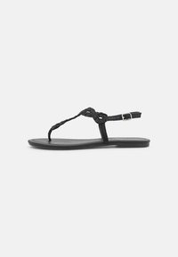 Sort flad sandal med flettet T-stropp og justerbar spænde ankelsnor, præsenteret på en ensfarvet hvid baggrund.
