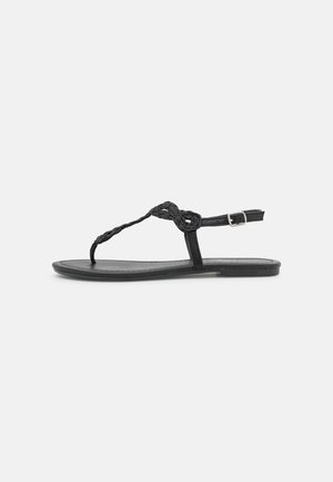 Sandale plate noire avec une sangle T tressée et une bride de cheville réglable à boucle, présentée sur un fond blanc uni.