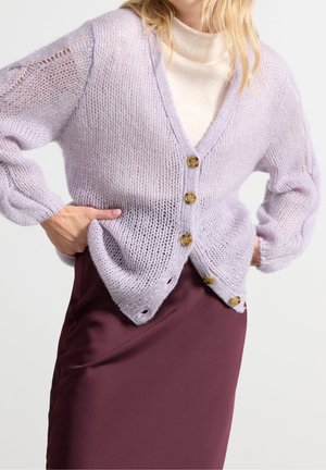 Cardigan lavorato a maglia di un leggero color lavanda con una vestibilità ampia, scollatura a V e tre bottoni di legno. Indossato sopra un dolcevita color crema e una gonna bordeaux.