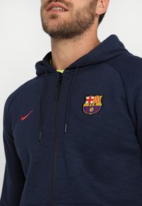 Felpa con zip di colore blu navale realizzata in tessuto testurizzato, con il logo rosso dello swoosh Nike e l'emblema del Barcellona FC sul lato sinistro del petto.