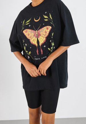 Person iført en sort oversized t-shirt med en stor farverig sommerfugl, blade, stjerner, halvmåne og teksten "TRUE. LOVE" på fronten.