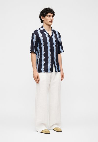 Άνδρας που φοράει κοντομάνικο μαύρο και μπλε κυματιστό ριγέ πουκάμισο, λευκό παντελόνι φαρδύ, και μπεζ παπούτσια, στέκεται μπροστά σε απλό φόντο.