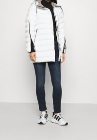 Vit vadderad pufferjacka med avtagbar huva, tillsammans med mörkblå jeans och sneakers i svart-vitt mönster. Slät och mjuk textur.