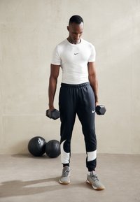 Pantalones de chándal negros de Nike con detalles en blanco, camiseta blanca ajustada, sosteniendo dos mancuernas negras, zapatillas grises. Entorno de gimnasio minimalista.
