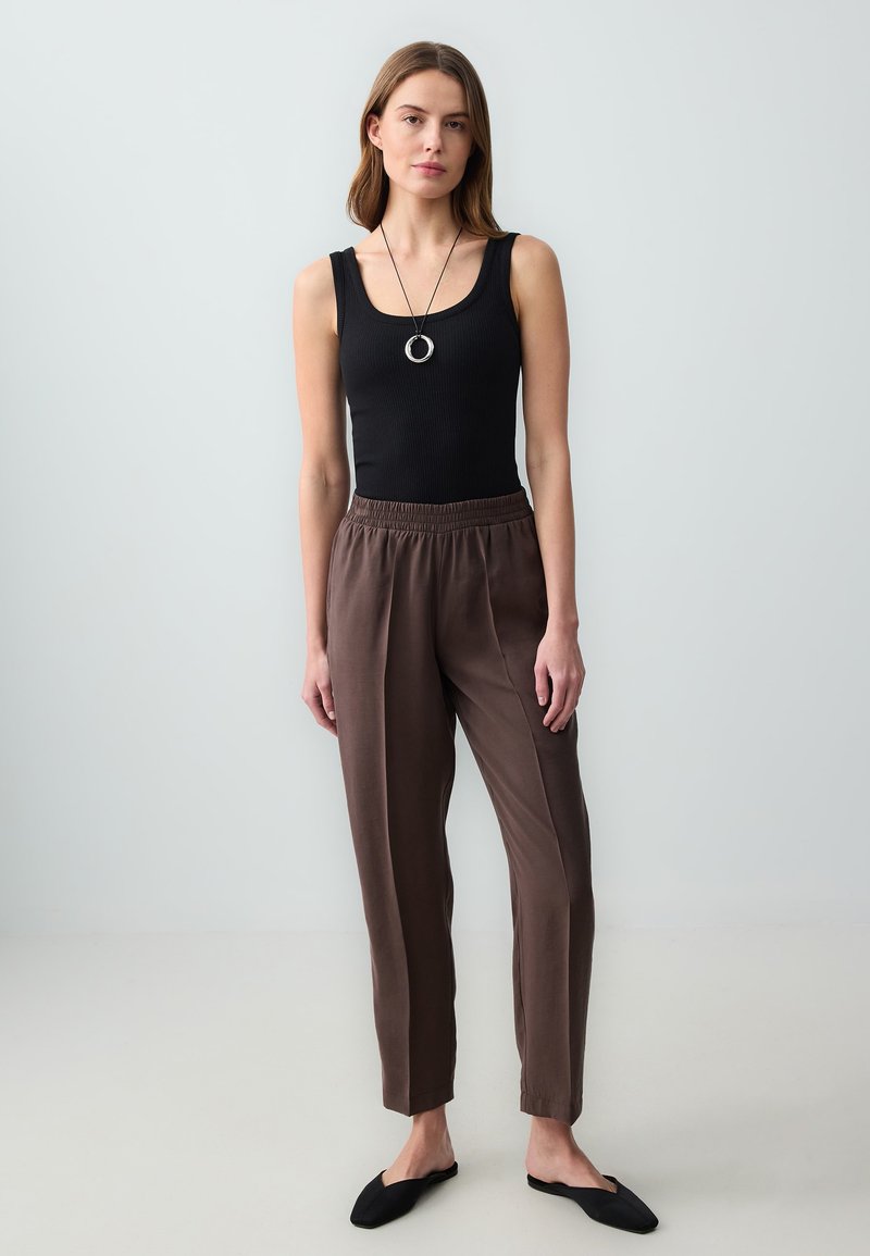 Femme debout portant un haut noir sans manches, un pantalon ample marron, des chaussures noires sans lacets, et un collier pendentif circulaire sur un fond uni.