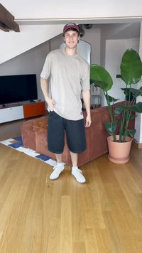 Lys grå t-skjorte, svarte shorts og hvite joggesko. Står i et rom med en plante, tregulv og en rød sofa i bakgrunnen.