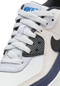 Tenisky Nike s bílým koženým svrškem a modrými a černými detaily, texturovaným černým panelem a bílými tkaničkami. Kulatý tvar špičky a vrstvený design.