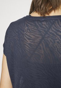 Námořnická modrá blůza s krátkým rukávem, která má průhledný, texturovaný zvířecí vzor. Zadní část má jemný design s hladkým povrchem.