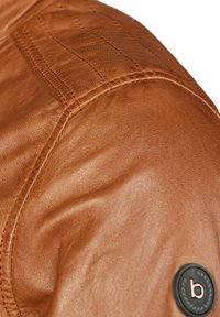 Acercamiento a una chaqueta de cuero marrón, que presenta una textura suave, detalles de costura en los hombros y un parche redondo con la letra "b."