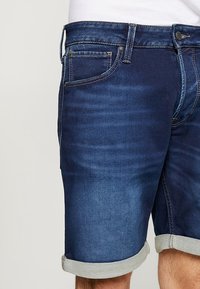 Farkkushortsit tummansinisessä sävyssä, joissa on käännehelma, kontrastiväriset ompeleet, etutaskut ja vetoketjulaskeuma napin sulkimella.