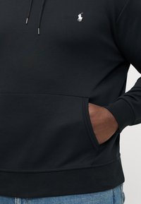 Svart hettegenser laget av mykt stoff med kengurulomme, ribbede mansjetter og en liten hvit logo på brystet. Avslappet passform.