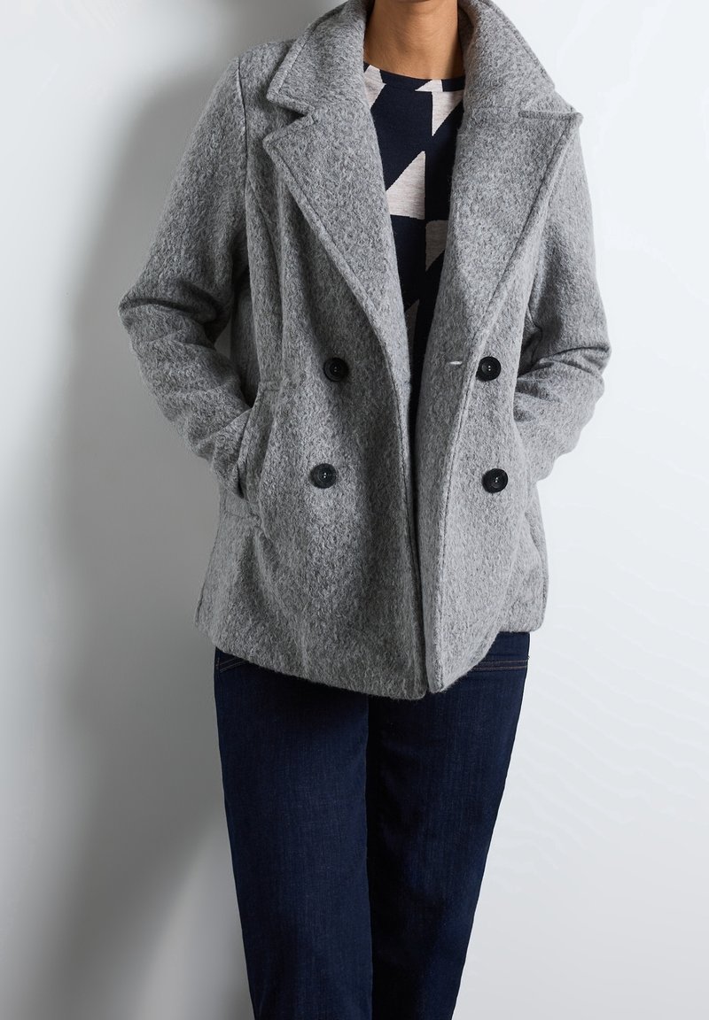 Personne portant un manteau en laine gris à double boutonnage avec des boutons noirs, un haut bleu marine à motif géométrique, et un jean bleu foncé, les mains dans les poches.