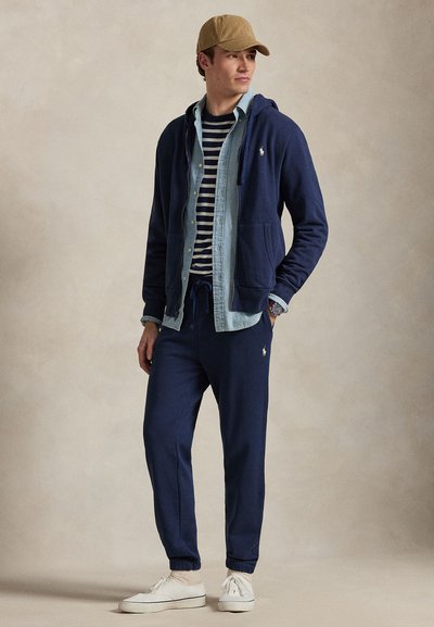 Tmavomodrá mikina s kapucňou a šnúrkami, vrstvená cez rifle a pruhované tričko. K tomu ladia tmavomodré tepláky a biele tenisky.