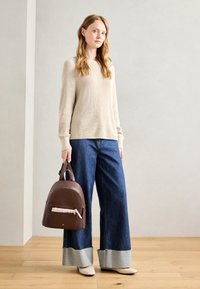 Maglione beige a costine, jeans in denim blu a gamba larga con orli sfrangiati, scarpe color crema e uno zaino in pelle marrone con dettagli floreali nella tasca.