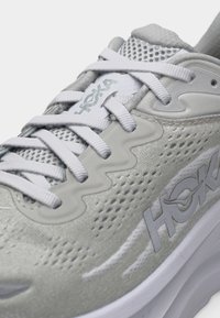 Κοντινό πλάνο ανοιχτόγκρι παπουτσιού τρεξίματος Hoka με λευκά κορδόνια, επάνω μέρος από δίχτυ και λογότυπο της μάρκας στη γλώσσα και στην πλάγια πλευρά.