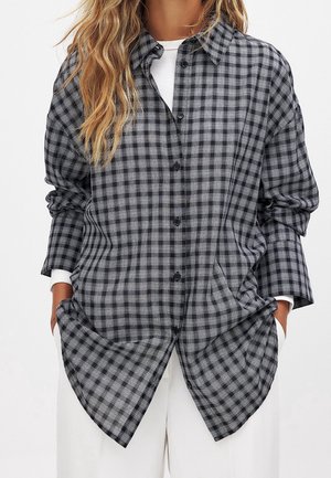 Femme portant une chemise oversize à carreaux noirs et gris boutonnée, par-dessus un haut blanc avec un pantalon blanc, les mains dans les poches.