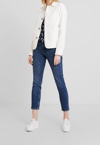 White corduroy jacket with button closure, paired with dark blue skinny jeans and white sneakers. The outfit features a floral print top underneath.