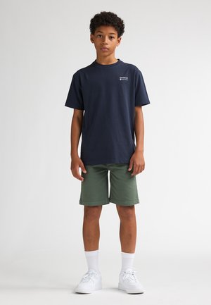 Tricou din bumbac bleumarin cu mâneci scurte, asortat cu pantaloni scurți verde măsliniu. Întregul outfit este completat de adidași albi și șosete albe până la gleznă.