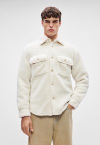 Hvit fleecejakke med myk tekstur, med knappestolpe foran og to brystlommer. Krage-design. Brukt over beige bukser.