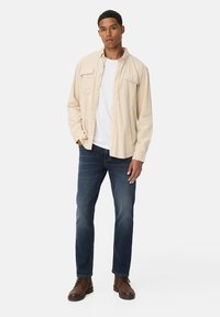 Chemise beige à manches longues avec des boutons sur un t-shirt blanc, portée avec un jean bleu foncé et des bottes marron à lacets, se tenant sur un fond blanc.