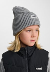 Grå stickad beanie med ribbad textur och vikbar mudd, med en rektangulär vit etikett med varumärket. Bärs med en svart väst.