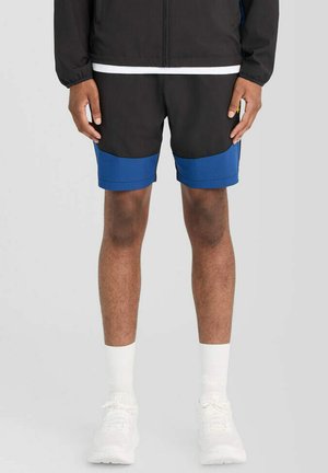 Man draagt zwart-blauwe colorblock korte broek, witte sokken, witte sneakers en een zwart jasje, staand tegen een effen achtergrond.